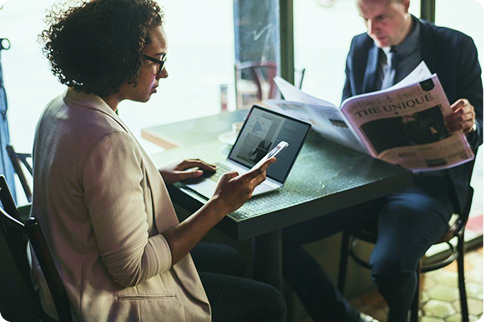 A woman uses a smartphone and laptop at a café table while a man in the background reads a newspaper.