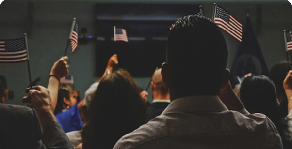 A group of people facing forward wave small American flags in an indoor setting, suggesting participation in a formal or patriotic event.
