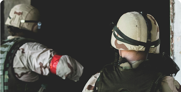 Two soldiers wearing camouflage uniforms and helmets stand near a dark doorway; one has a red armband.