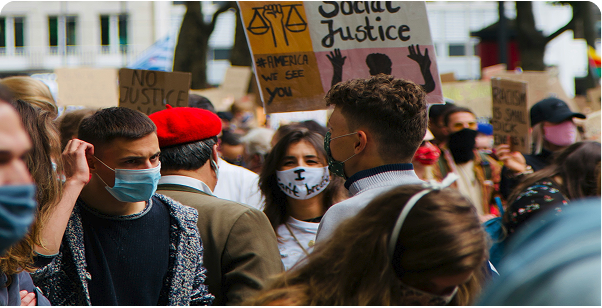A group of people wearing face masks gather outdoors, holding signs with messages about justice and social issues.