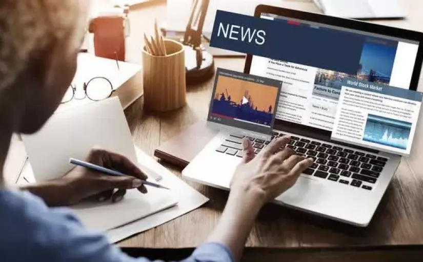 A person types on a laptop displaying news articles and graphs while taking notes on paper in an office setting.
