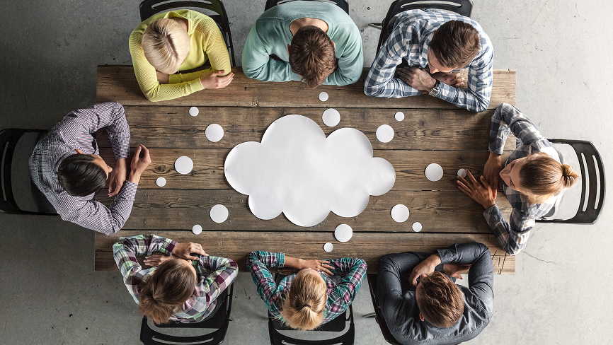 Eight people sit around a wooden table viewed from above, with a large white cloud-shaped cutout and several round paper circles on the table.
