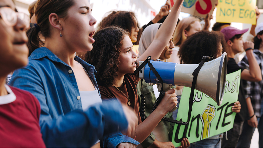 A group of diverse young people at a protest, holding signs and a megaphone, demonstrating together outdoors.