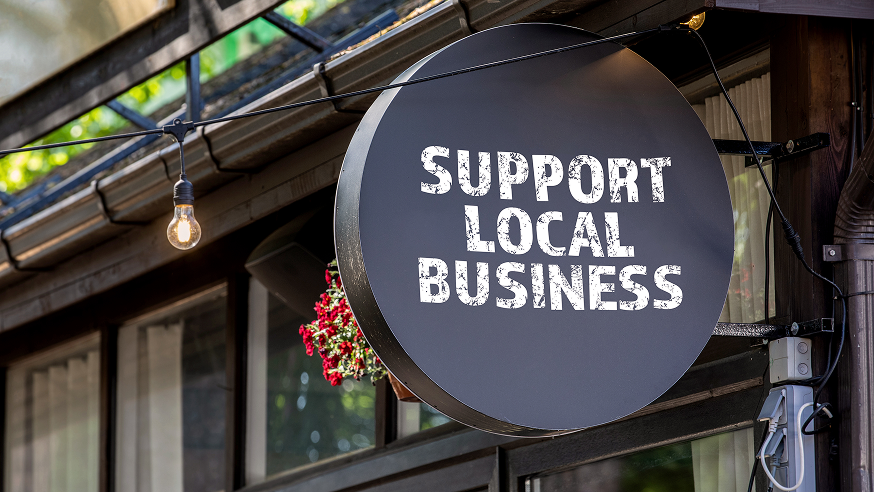 A round sign reads "Support Local Business" in bold white letters, hanging outside a building with a string light and some red flowers visible in the background.