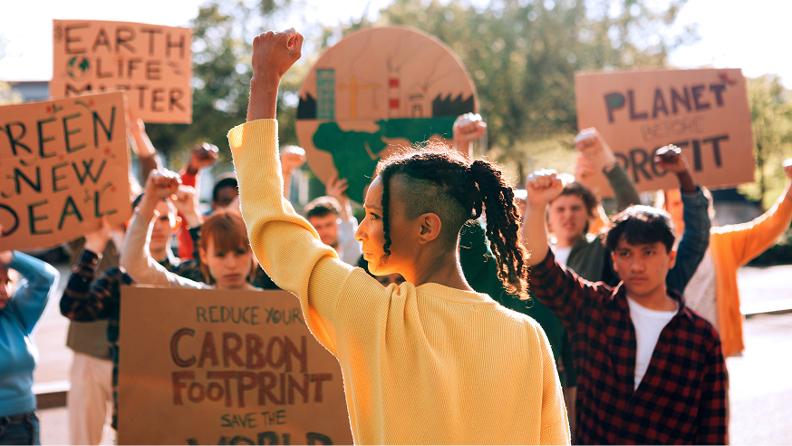 A group of people hold protest signs and raise their fists at an outdoor climate change rally. Signs reference topics like carbon footprint, profit, and the Green New Deal.