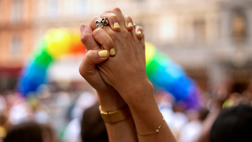 Two hands with yellow nail polish are clasped together in front of a blurred rainbow-colored arch at an outdoor event.