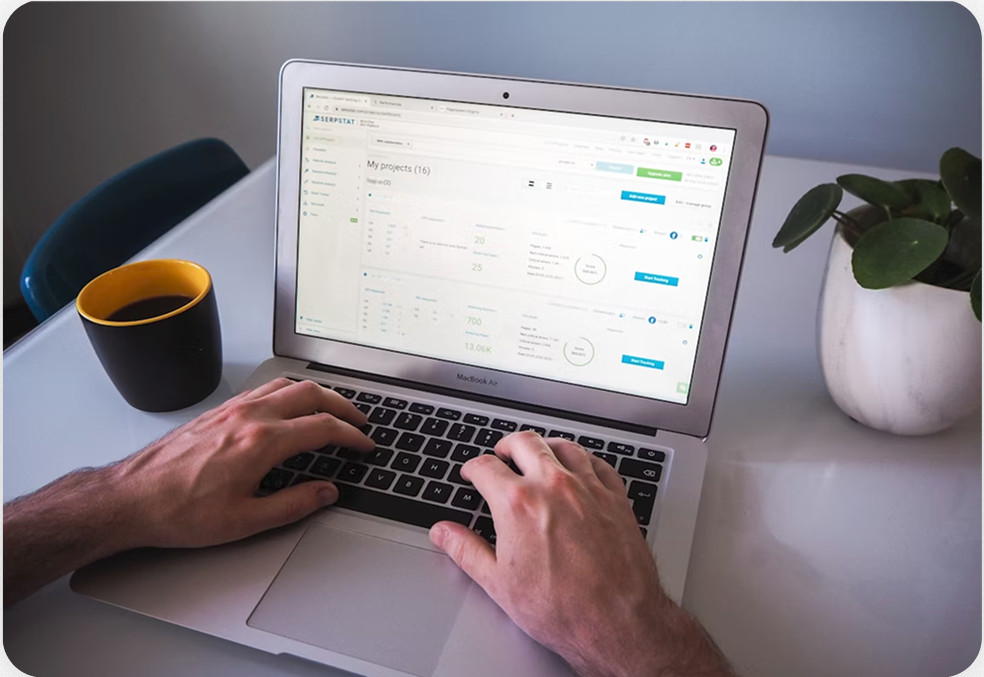 Person working on a laptop displaying a project management dashboard, with a cup of coffee and a potted plant on the white desk.