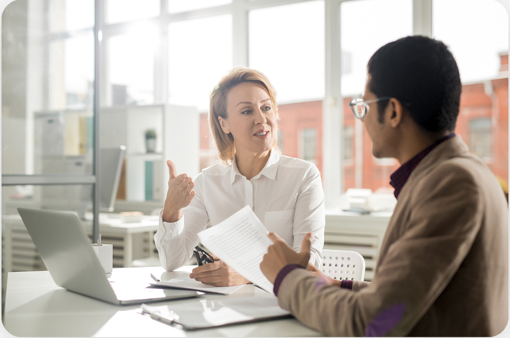 Two people sit at a desk in an office, engaged in a conversation. One holds documents, while the other gestures with her hand. A laptop is open on the table.
